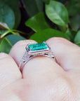 Ring with natural emerald on a finger against a leafy background.