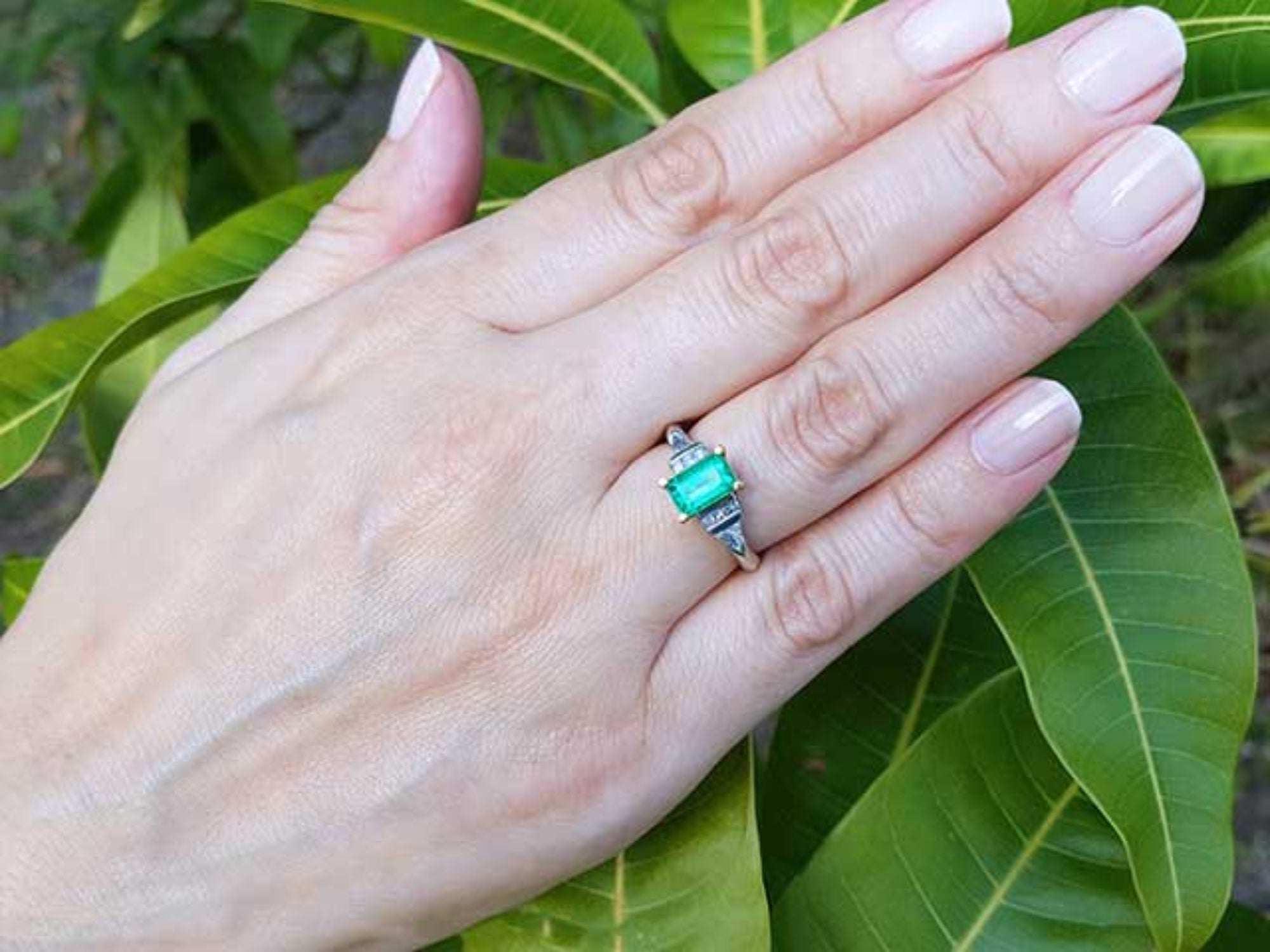 Hand wearing a natural emerald ring with leaves in the background.