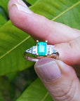 A platinum emerald ring being held between fingers against a leafy background.