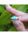 A platinum emerald ring being held between fingers against a leafy background.