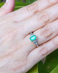 Hand wearing a ring with natural emerald against a leafy background.