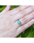 Hand wearing a ring with natural emerald against a leafy background.