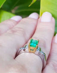 Gold and platinum ring with a natural emerald on a hand against a green leafy background.