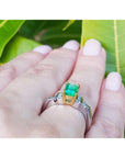 Gold and platinum ring with a natural emerald on a hand against a green leafy background.
