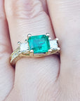 This is a women's emerald ring with princess cut side diamonds on a finger.