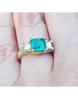 This is a women's emerald ring with princess cut side diamonds on a finger.