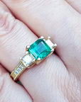 Bridal's engagement ring with emerald and side diamonds princess cut on a finger.