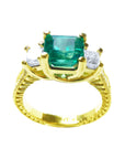 A 18k yellow gold emerald ring with side princess cut diamonds, on a white background.