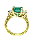 A lateral profile view including gallery of an emerald ring with side princess cut diamonds in 18k yellow gold.