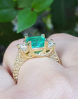 A Brial's may birthstone emerald ring held in a hand against a blurred green leafy background.