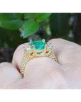 A Brial's may birthstone emerald ring held in a hand against a blurred green leafy background.