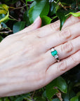Hand wearing a ring with emerald stone and side diamonds against a green leafy background.