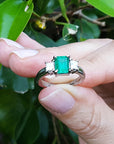 Hand holding a natural Colombian emerald ring with side princess cut diamonds against a leafy background.