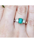 This three stone natural colombian emerald ring with side princess cut diamonds is on a finger.