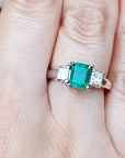 This three stone natural emerald ring with side princess cut diamonds is on a finger.
