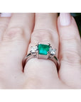 18k white gold three-stone ring with a vibrant emerald and two princess-cut diamonds on a hand.