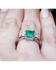 18k white gold three-stone ring with a vibrant emerald and two princess-cut diamonds on a hand.