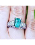 A women's Colombian emerald ring in 18k white gold and accent diamonds on a finger.