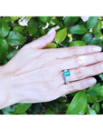 Hand wearing a ring with a natural Colombian emerald against a green leafy background.