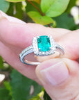 Emerald ring with diamond accents held between fingers against a green leafy background.