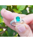 Emerald ring with diamond accents held between fingers against a green leafy background.