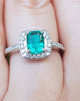 A women's emerald ring cushion cut with halo diamond on a finger.