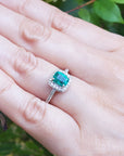 Hand wearing a cushion cut emerald ring against a natural background.