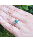 Hand wearing a cushion cut emerald ring against a natural background.