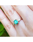 A gold ring with emerald and side baguette diamonds on a hand against a blurred green background.