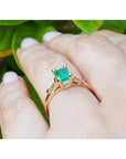 Yellow gold ring with emerald stone on a hand against a green leafy background.