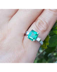An emerald ring with side diamonds in white gold on a finger with a natural background.