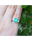 Emerald and side diamonds ring on a finger with a green leafy background.