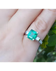 A GIA certified natural colombian emerald ring with side princess cut diamonds in 14k white gold on a finger.