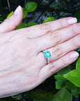 Hand wearing a ring with an emerald stone against a green leafy background.