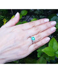 Hand wearing a ring with an emerald stone against a green leafy background.