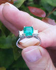 Natural Colombian emerald rings held between fingers against a blurred natural background.