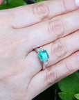 Hand wearing an emerald ring with side diamonds against a green leafy background.
