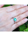 Hand wearing an emerald ring with side diamonds against a green leafy background.
