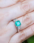 Emerald ring with diamond accents on a hand against a natural background.