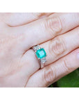 Emerald ring with diamond accents on a hand against a natural background.