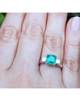Square emerald-cut colombian emerald ring three stone with side princess cut natural diamonds on a finger.