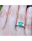 Square emerald-cut colombian emerald ring three stone with side princess cut natural diamonds on a finger.