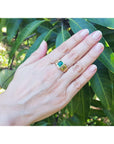 Hand wearing a gold mens ring with a natural colombian emerald against a leafy background.