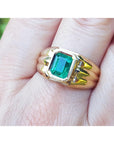 Gold mens solitaire ring with a rectangular bezel set emerald on a hand.