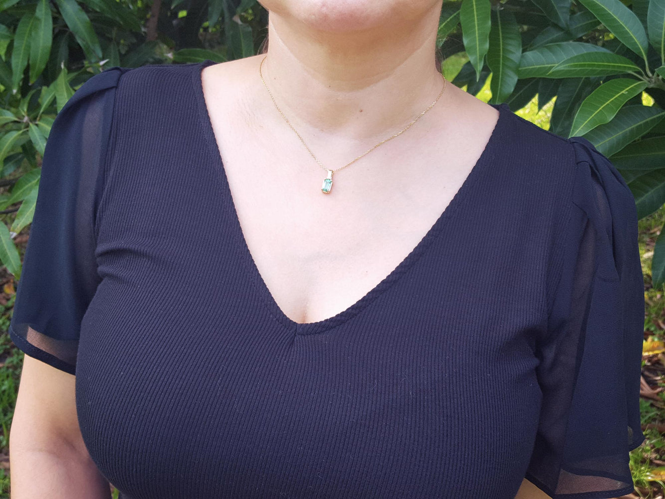 A woman wearing a dark blue top with sheer sleeves and an emerald solitaire pendant against a green leafy background.