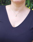 A woman wearing a dark blue top with sheer sleeves and an emerald solitaire pendant against a green leafy background.