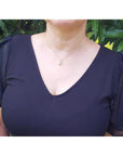 A woman wearing a dark blue top with sheer sleeves and an emerald solitaire pendant against a green leafy background.