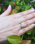 Hand wearing a solitaire emerald ring bezel set in yellow gold against a green leafy background