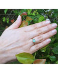 Hand wearing a 0.81 ct. medium bluish green emerald solitaire ring, bezel set in 14K yellow gold.