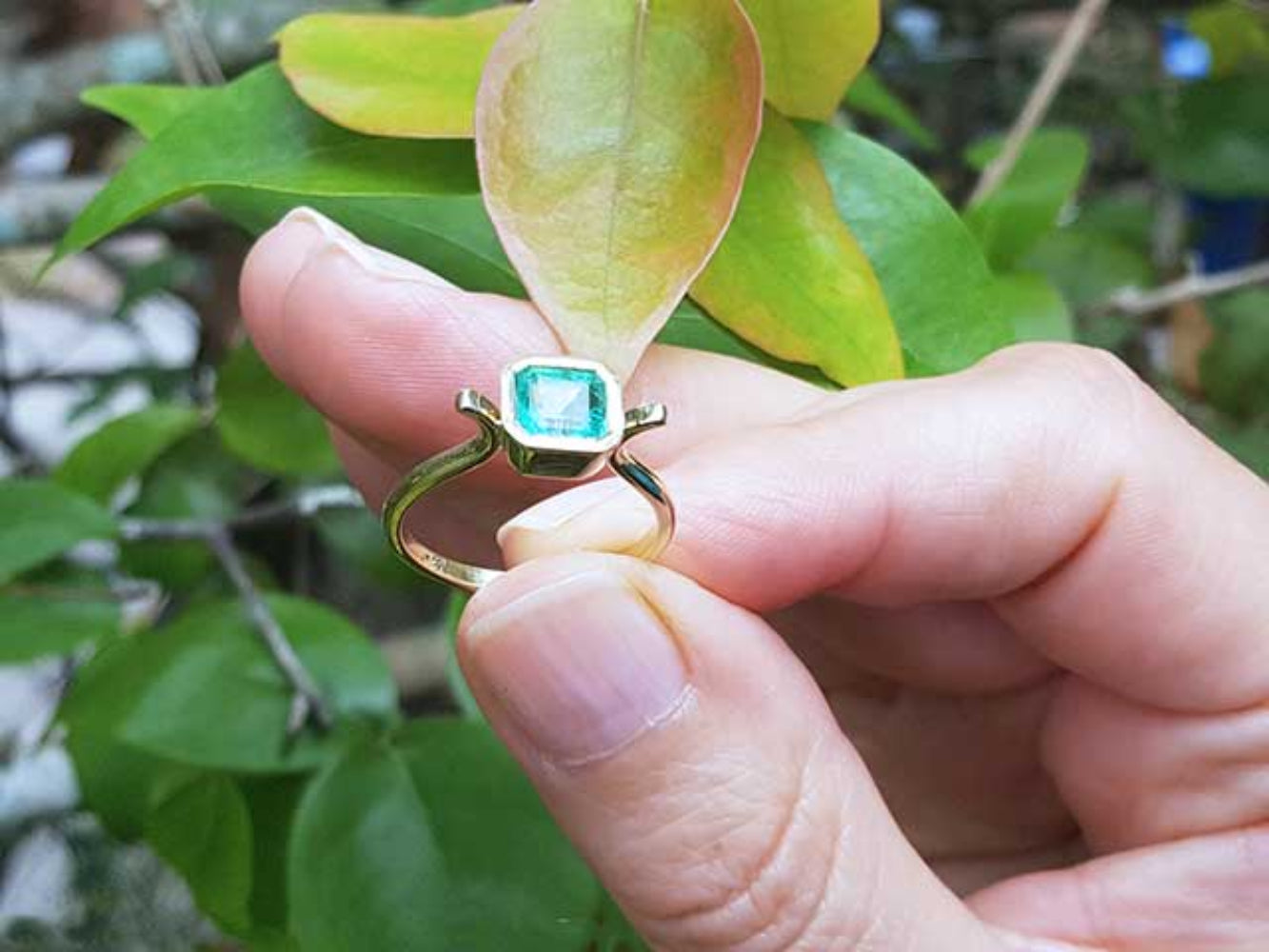 A solitaire emerald ring held between fingers against a leafy background